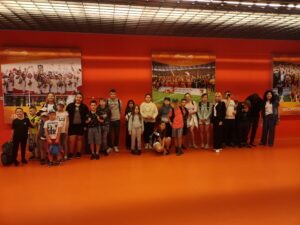 Grupa dzieci i dorosłych pozuje do zdjęcia na pomarańczowym tle, przed wielkimi plakatami przedstawiającymi sceny z meczów piłkarskich na stadionie miejskim, w tym radosnych zawodników z trofeami.