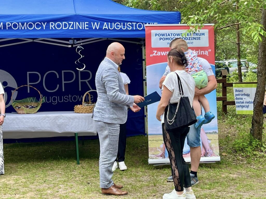 Zdjęcie z wydarzenia organizowanego przez Powiatowe Centrum Pomocy Rodzinie w Augustowie. Mężczyzna w jasnoszarym garniturze wręcza dokument kobiecie, obok stoi mężczyzna trzymający małe dziecko. W tle namiot z logo PCPR i baner z hasłem 'Zawsze z rodziną'. Na drzewie widoczny plakat z napisem 'Dzieci potrzebują domu – zostań rodziną zastępczą'. Wokół drzewa i samochody zaparkowane w tle.
