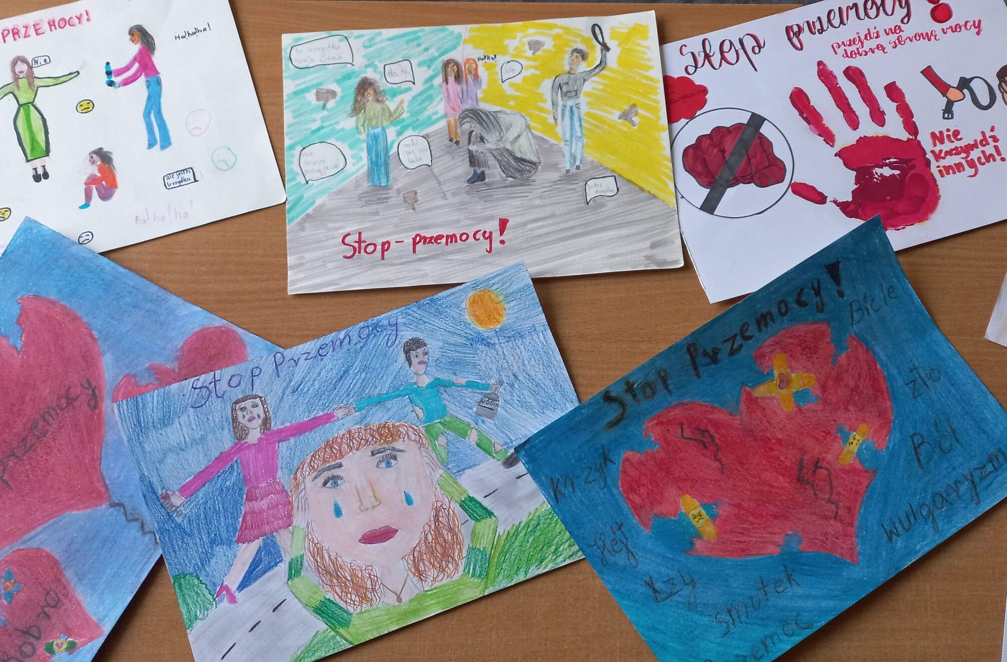 Na zdjęciu widać dziecięce rysunki o tematyce „Stop przemocy”, ułożone na drewnianym blacie. Dominują czerwone serca, łzy, postacie broniące innych i hasła przeciw agresji. Niektóre prace symboliczne, inne przedstawiają sceny przemocy fizycznej lub psychicznej.
