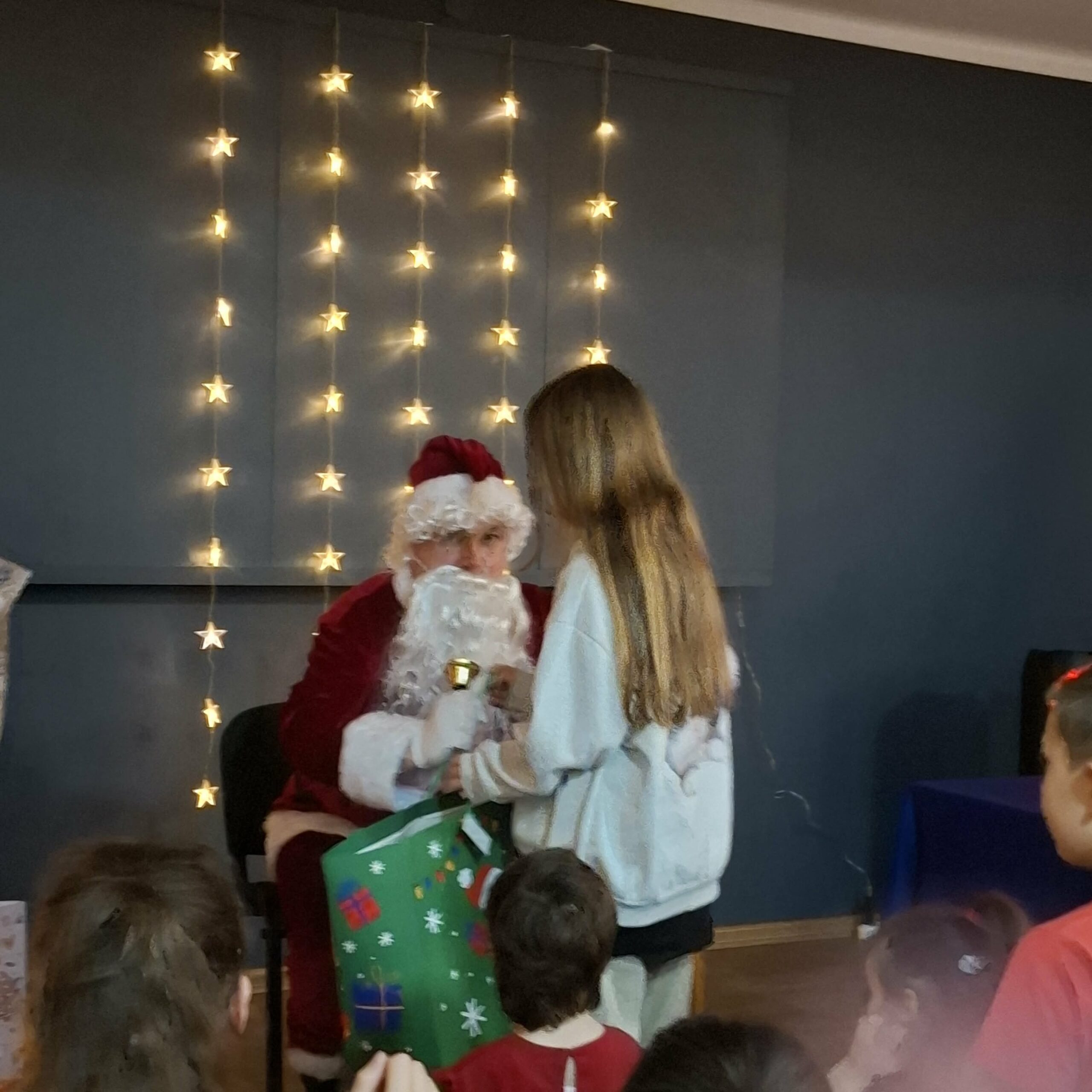 Dziewczynka odbiera prezent od Świętego Mikołaja, który siedzi wśród dekoracji ze światełkami. Obok niego stoi duża torba z prezentami i kilka dzieci obserwujących sytuację.